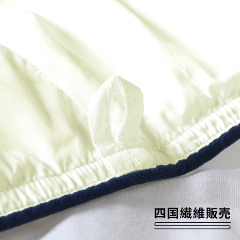 米白色雲絨被特寫，露出邊緣的深藍色滾邊和一個固定環，被子表面有細緻的縫線和絎縫紋理，材質看起來柔軟蓬鬆。