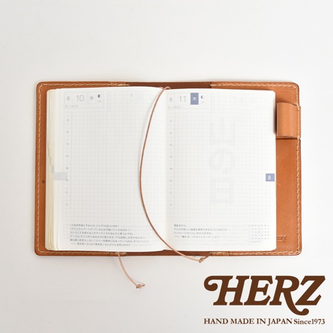 HERZ 品牌標誌，下方為 HAND MADE IN JAPAN Since1973 字樣，圖片中央為一本打開的棕色皮革手帳，內頁為日曆格式，以俯視角度拍攝。