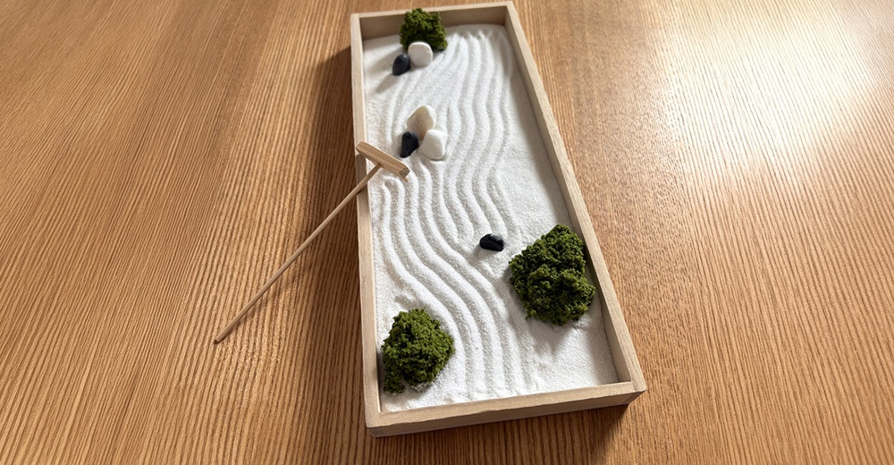 一個長方形的木製托盤擺放在木質桌面上，盤中鋪滿白色細沙並刻畫出流線型的波紋，點綴著仿真青苔、數顆圓潤的小石子以及一把精緻的木製耙子，展現日式枯山水的靜謐意境。