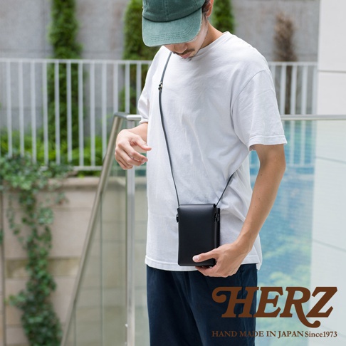 一名男子身著白色T恤和深藍色長褲，斜挎一個黑色皮革手機包。手機包為長方形，以細長黑色皮革背帶連接，包蓋處有磁扣設計，整體為簡約風格。