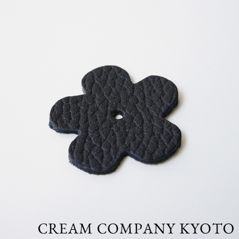 京都職人手作的黑色皮革花朵造型吊飾，表面有細緻紋理，中心有一個圓形孔洞，可用於收納包的裝飾或配件。