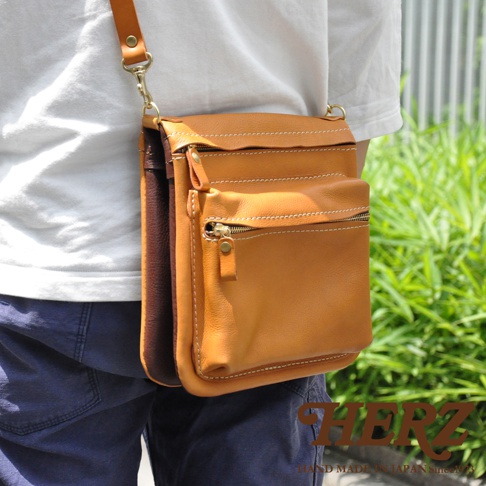 職人手作皮革翻轉雙色直式斜背包，主體為咖其色皮革，搭配深棕色皮革側邊，正面有兩個拉鍊口袋，整體風格簡約實用。