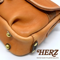 職人手作皮革鹿角扣繩郵差包，呈現原色皮革的自然紋理與光澤，包身設有金屬圓釘裝飾，搭配深棕色皮革標籤，展現簡約復古風格。