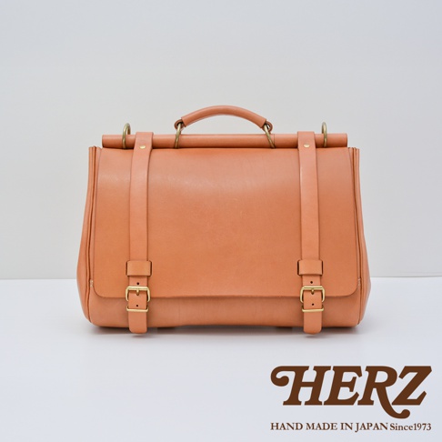 一個褐色的皮革側背包，附有金色扣環和肩帶，包包上方有一個把手，下方印有「HERZ」的品牌標誌和「HAND MADE IN JAPAN Since1973」的文字。