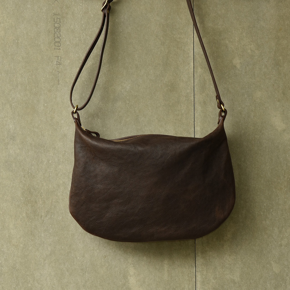 深棕色皮革製成的單肩半月包，帶有拉鍊開口和可調節的皮革肩帶，以金屬環連接。