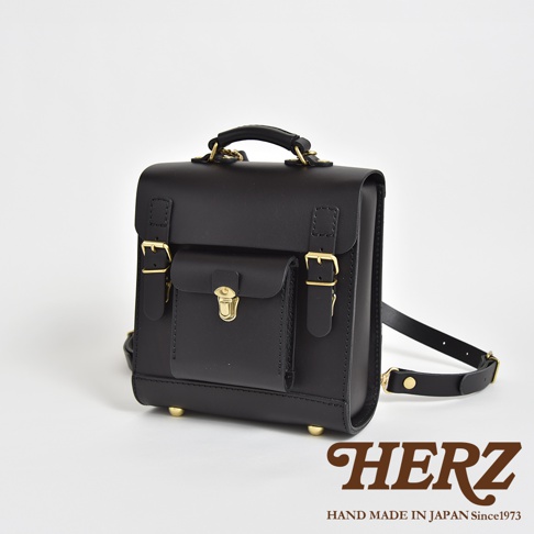 職人手作黑色皮革經典學院風迷你後背包，正面有翻蓋式口袋，搭配金色鎖扣及調節扣，底部有四個金屬腳釘。