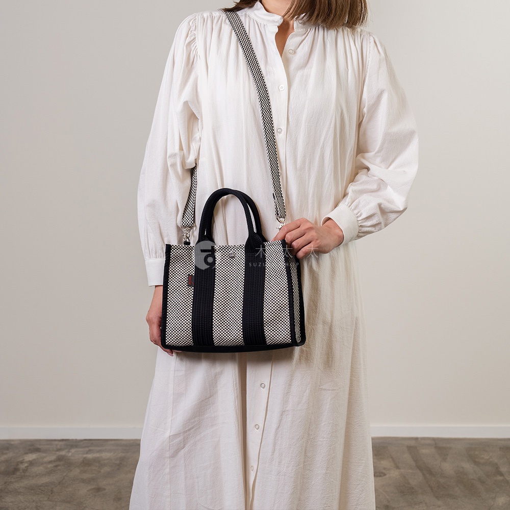 一位身穿白色長洋裝的女子，肩背一個黑白條紋、方形的刺子織托特包，包包上有提把和可拆卸的斜背帶。