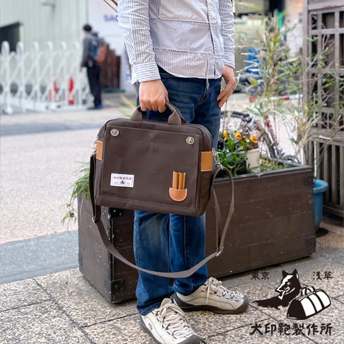 深棕色純棉帆布三用橫式側背包，配有棕色皮革飾邊和手提把，以及可拆卸的肩帶，包包上有品牌標誌貼布。