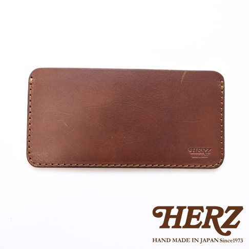 日本職人品牌 HERZ 手作皮革紙鈔收納夾，採用棕色軟皮革製成，表面呈現自然的皮革紋理，邊緣以對比色的粗縫線手工縫製，右下角壓印有 HERZ 品牌標誌與產地說明，整體設計簡約精緻。