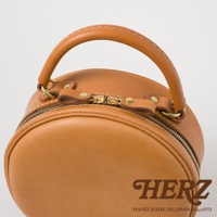 HERZ 品牌標誌與 Since 1973 字樣，搭配手工皮革圓形側背包的提把及拉鍊細節