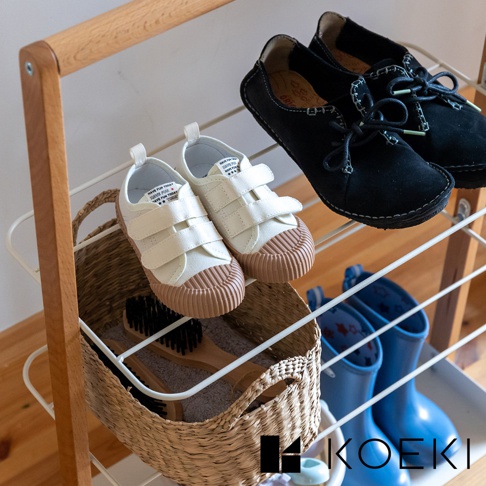 木質框架鞋架上，白色魔鬼氈帆布童鞋與黑色麂皮綁帶平底鞋，下方籃子內有兩支木柄刷。