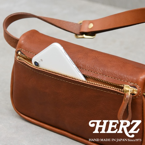 職人手作皮革Organ斜背包，棕色皮革材質，拉鍊開口露出部分銀色手機，可調節的皮革背帶配有金色扣環。