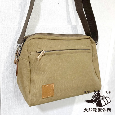 一個卡其色石洗帆布旅行側背包，配有深棕色帆布背帶，包包前方有一個拉鍊口袋，下方有一個方形皮革標誌，右下方有狗和行李的黑色圖案，以及日文文字。