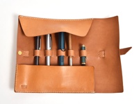 職人手作淺棕色皮革捲捲收納筆袋，內有五個筆槽，分別裝有黑色、銀色、藍色、木質和綠色鋼筆。筆袋以白色縫線裝飾，並以黃銅色鈕扣和皮革帶固定。