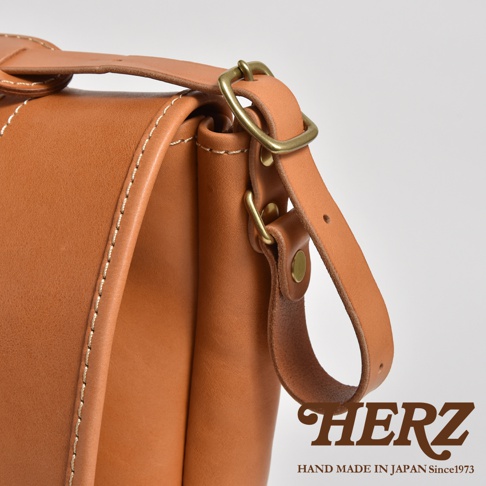 職人手作皮革復古郵差包，採用淺棕色皮革，搭配黃銅色金屬扣環與鉚釘，包款縫線清晰，展現紮實工藝。