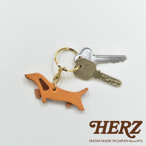 職人手作皮革狗狗造型鑰匙圈，淺棕色皮革製成，搭配深棕色耳朵與腳部細節，附金色鑰匙圈與掛鉤，與兩把銀色鑰匙一同展示。