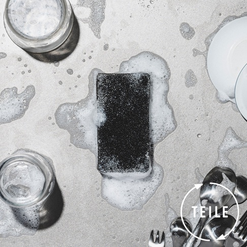 黑色超細纖維清潔海綿，置於充滿泡沫的淺色背景上，周圍有玻璃杯、白色盤子和銀色餐具。