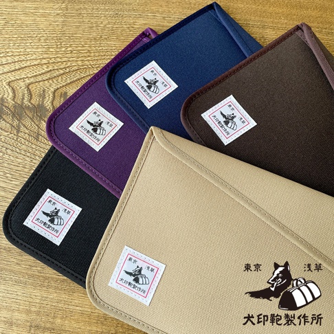 犬印純綿帆布平板收納包，展示了黑色、紫色、深藍色、棕色和卡其色五種顏色。每個收納包皆有印有「犬印製作所」標誌的白色方形標籤。