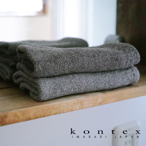 兩條深灰色針織紋純棉毛巾堆疊在木架上，左側有毛巾的鏡面反射。