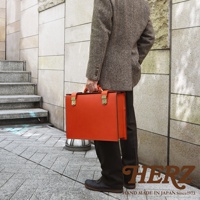 職人手作皮革機師箱 BC-31，亮橘色皮革，配有金色鎖扣和提把。箱體方正，線條簡潔，展現經典設計。