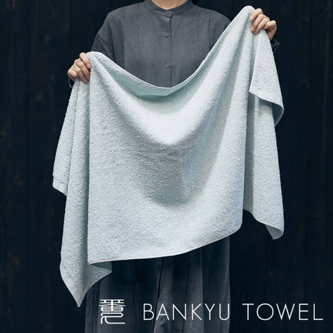 淺藍色長纖維浴巾，質地柔軟，表面有細緻的絨毛紋理，由兩人雙手展開，背景為深色木紋。