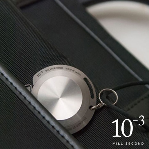 銀色鋁製卷尺，圓形錶盤，上面刻有「10-3 MILLISECOND MADE IN JAPAN」字樣，附有鑰匙圈和黑色掛繩，放置於黑色織物表面。