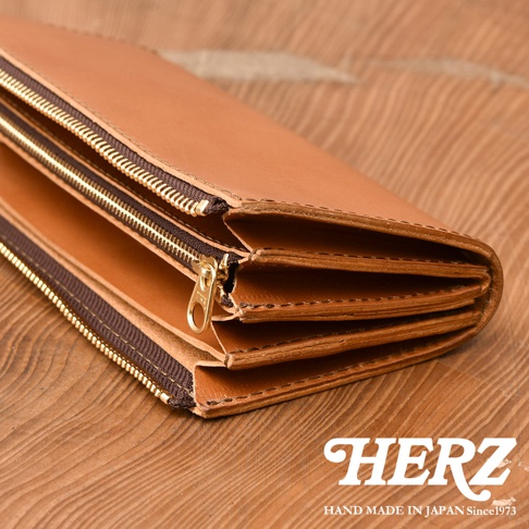 日本職人手工製作的HERZ皮革長夾，呈現側面的多層結構與金色拉鍊細節，採用優質Dakoda皮革製成，展現溫潤的棕色質感與細緻的縫線工藝，放置於木紋桌面上，右下角印有HERZ品牌標誌。