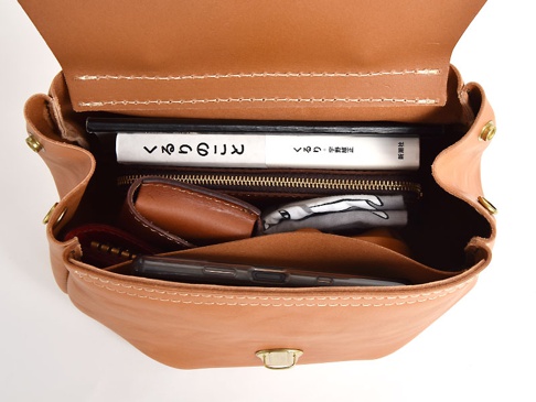 職人手作皮革屋瓦風迷你後背包 (R-6)，以天然皮革製成，呈現溫潤的焦糖棕色。包內可見書本、手機、零錢包及一條印花布，展示其收納空間。包身有金屬扣環及白色車縫線裝飾。