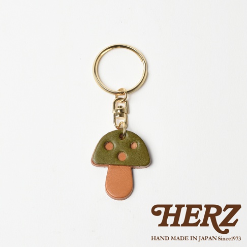 職人手作皮革香菇造型鑰匙圈，傘蓋為綠色皮革，帶有三個橘色圓點裝飾，傘柄為棕色皮革。搭配金色鑰匙圈和掛鉤，整體呈現可愛的蘑菇造型。
