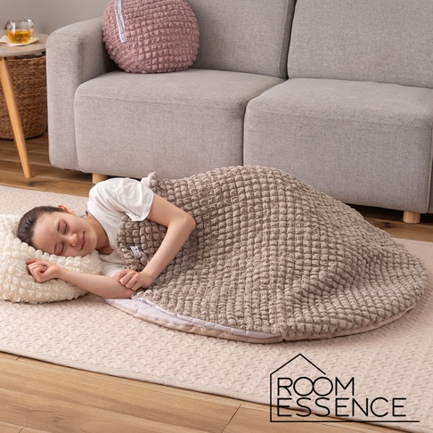 一位女性仰臥在米色地毯上，蓋著一個淺棕色、表面有蜂窩狀紋理的夾心包覆圓墊。她頭枕著一個奶油色紋理枕頭，閉著眼睛，表情放鬆。背景是淺灰色的沙發和旁邊的小桌子。