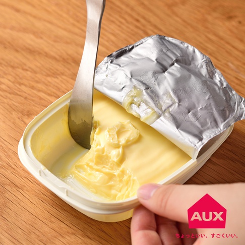 一個銀色金屬抹刀正在從一個裝有黃色奶油的白色塑膠容器中抹取奶油。容器被鋁箔蓋部分覆蓋，鋁箔上有壓紋。木質背景。