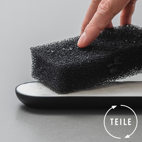 一隻手正拿著黑色網狀海綿，海綿上方有細小的水珠，下方是一個帶有白色內襯的黑色長方形皂盤，背景為淺灰色。