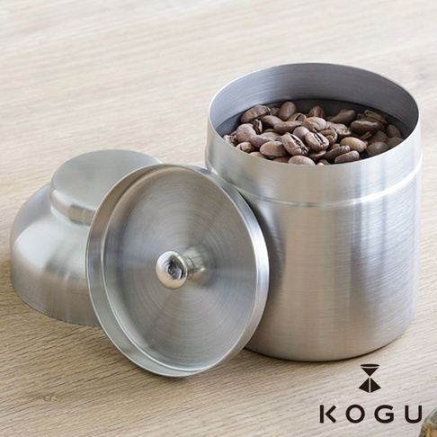 日製18-8不鏽鋼儲豆罐，罐身裝有咖啡豆，附帶一個銀色不鏽鋼量匙，置於木質桌面。