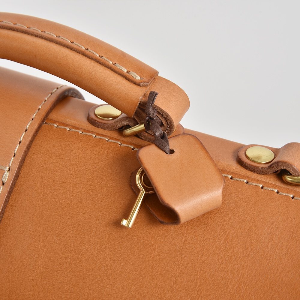 棕色皮革公事包的特寫，展示了手柄、黃銅五金件、縫線細節和一個帶有小鎖的皮革吊牌。