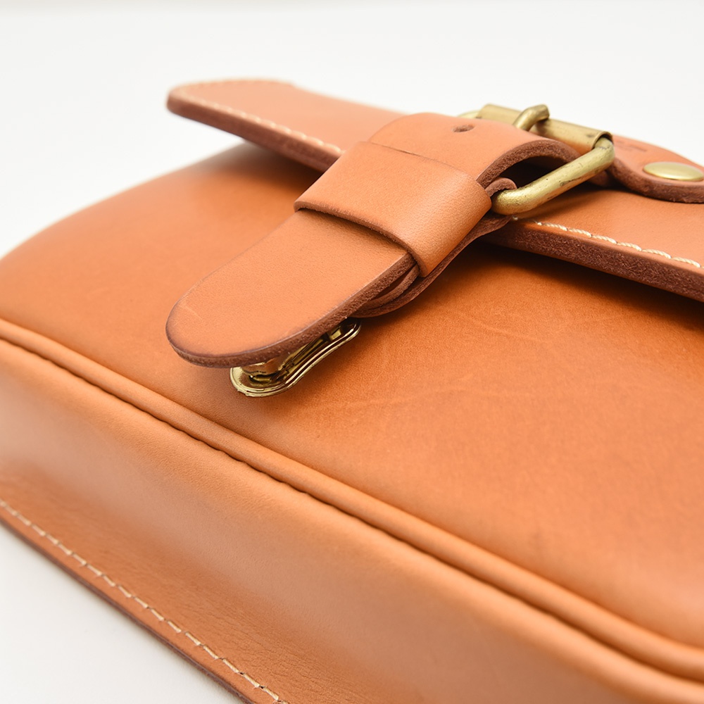 職人手作皮革單扣經典斜背包，採用光滑皮革製成，呈現自然色澤，搭配復古黃銅單扣和金屬鉤環，細膩車線展現職人手工質感。