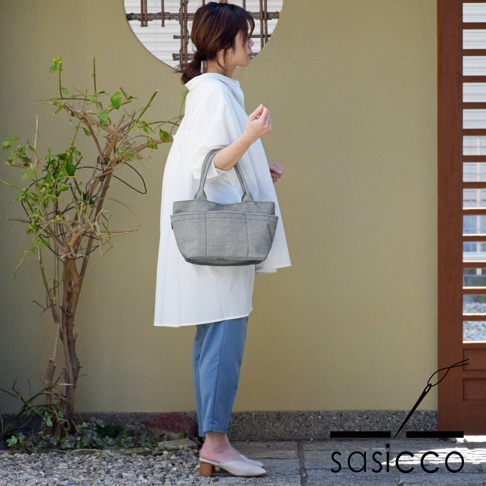 一位女性身穿白色寬鬆上衣和藍色長褲，手提一個灰色三河木綿刺子織小巧托特包，包包有側袋和提把，整體風格簡約。