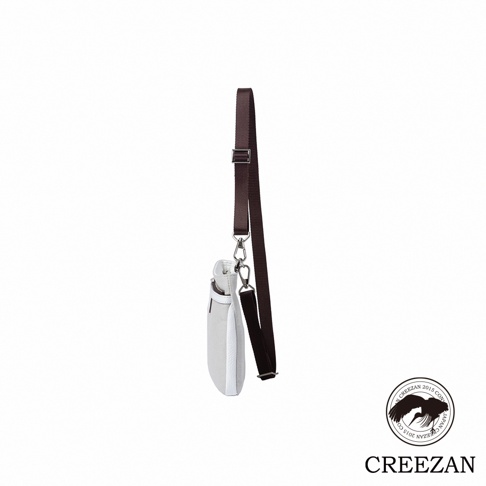 這是一個側面視角的 CREEZAN 品牌隨身斜背包，主體採用淺灰色義大利帆布搭配白色皮革飾邊，配有深褐色可調節式肩帶與金屬掛扣，右下角印有該品牌經典的鶴形圖案商標。