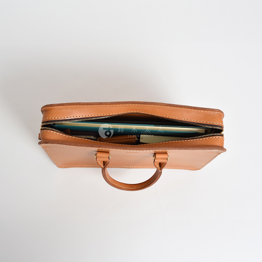 職人手作天然棕色皮革公事包，頂部拉鍊開啟，露出包內裝有書籍和皮夾。包身有縫線細節與兩個手提把。