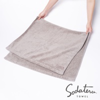 一件淺灰色、蓬鬆柔軟的棉質浴巾，表面有細緻的絨毛觸感，角落處有一個印有品牌名稱的標籤。