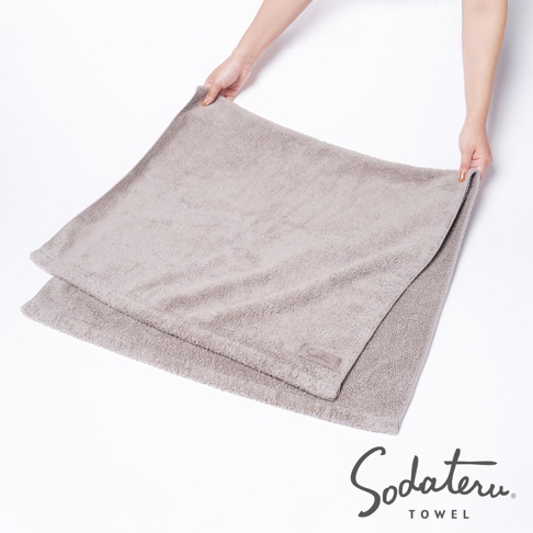 一件淺灰色、蓬鬆柔軟的棉質浴巾，表面有細緻的絨毛觸感，角落處有一個印有品牌名稱的標籤。