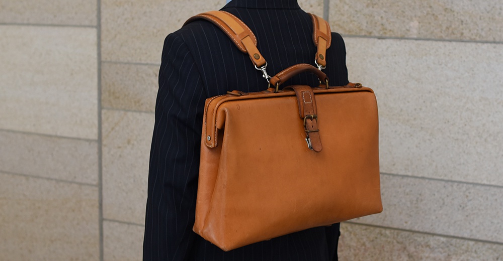 棕色皮革軟式杜勒斯包，配有肩帶和提把，包身有皮帶扣設計，由人體背部視角呈現。