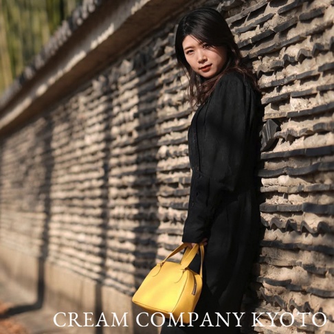 一位女子手持亮黃色半月形迷你手提包，包款為皮革材質，搭配黑色長版洋裝，背景為古樸的磚牆。