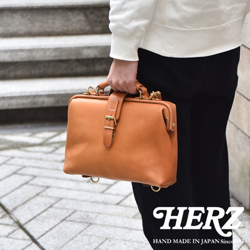 職人手作皮革迷你軟皮杜勒斯包，採用光澤感鞣製皮革製作，呈現溫潤的焦糖棕色，搭配金色金屬扣環與提把，設計簡約復古。