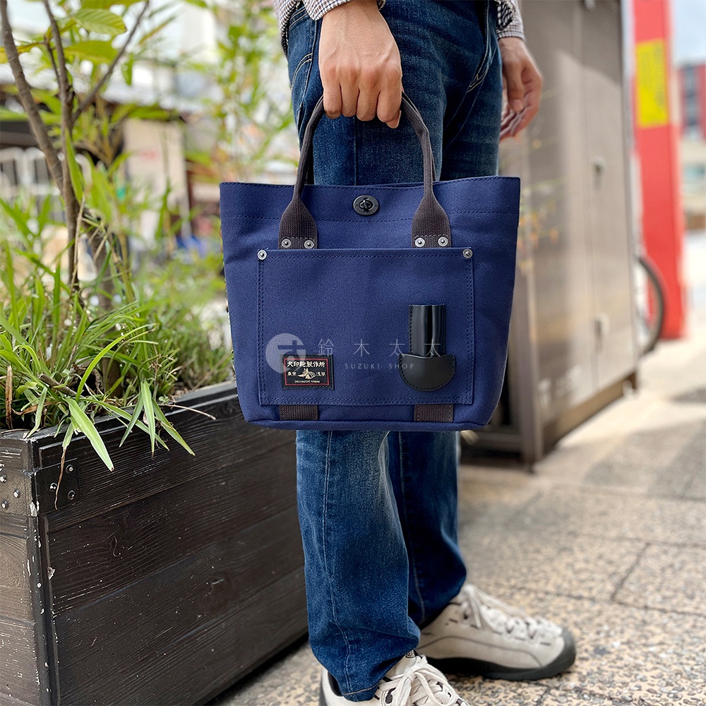 深藍色帆布水桶托特包，配有黑色皮革提把與細節，正面有標籤及一個黑色皮革套。