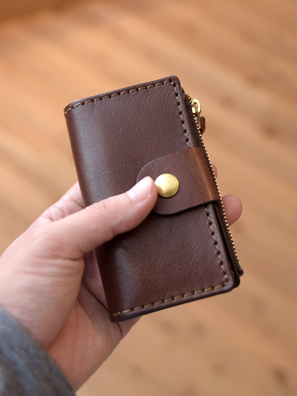一個由棕色皮革製成的職人手作零錢鑰匙包，配有金色拉鍊和按扣，以及明顯的縫線。