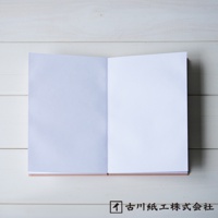 一本打開的和紙御朱印帳，左頁為淡紫色，右頁為白色。封面為粉紅色，攤開放置於淺色木紋背景上。