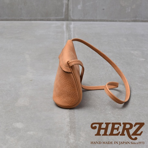 一個淺棕色皮質的斜背包，以側面角度展示，包包的皮質紋理清晰可見，配有細長的皮質背帶。背景為灰色水泥地面，右下角印有HERZ字樣和HAND MADE IN JAPAN Since1973的文字。