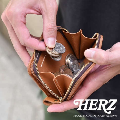 雙手握持一個日本HERZ手工製作的褐色皮革雙折錢包，內部空間寬敞，正展示從隔層中取出幾枚硬幣的細節，皮夾邊緣以精緻的拉鍊環繞，整體呈現出職人手工打造的質感。