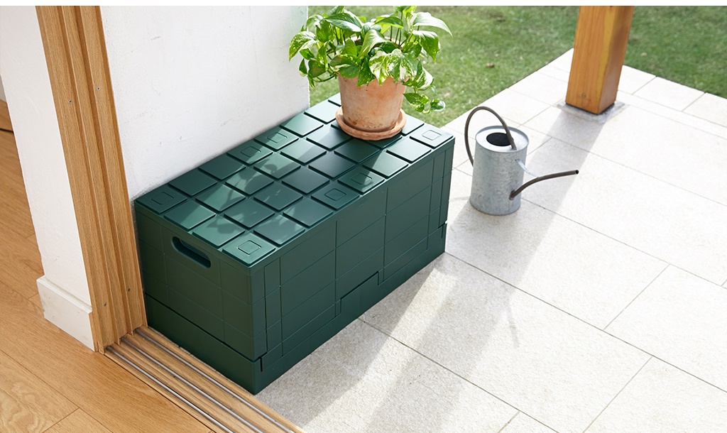 深綠色 Grid 格子磚可堆疊摺疊收納椅，頂部有菱形凹槽設計，側邊有提把，材質為塑膠，放置於戶外淺色磁磚地面上。