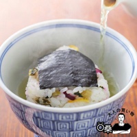 碗中盛著白米飯飯糰，飯糰上覆蓋著深綠色海苔片，旁邊有漬物點綴，碗外緣有藍色邊框，整體呈現日式風格，由上方傾倒著茶湯。